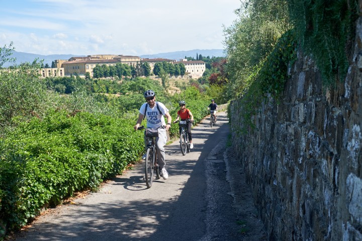 Three cyclists ride on a narrow path beside a stone wall with a scenic background of trees and a large building.