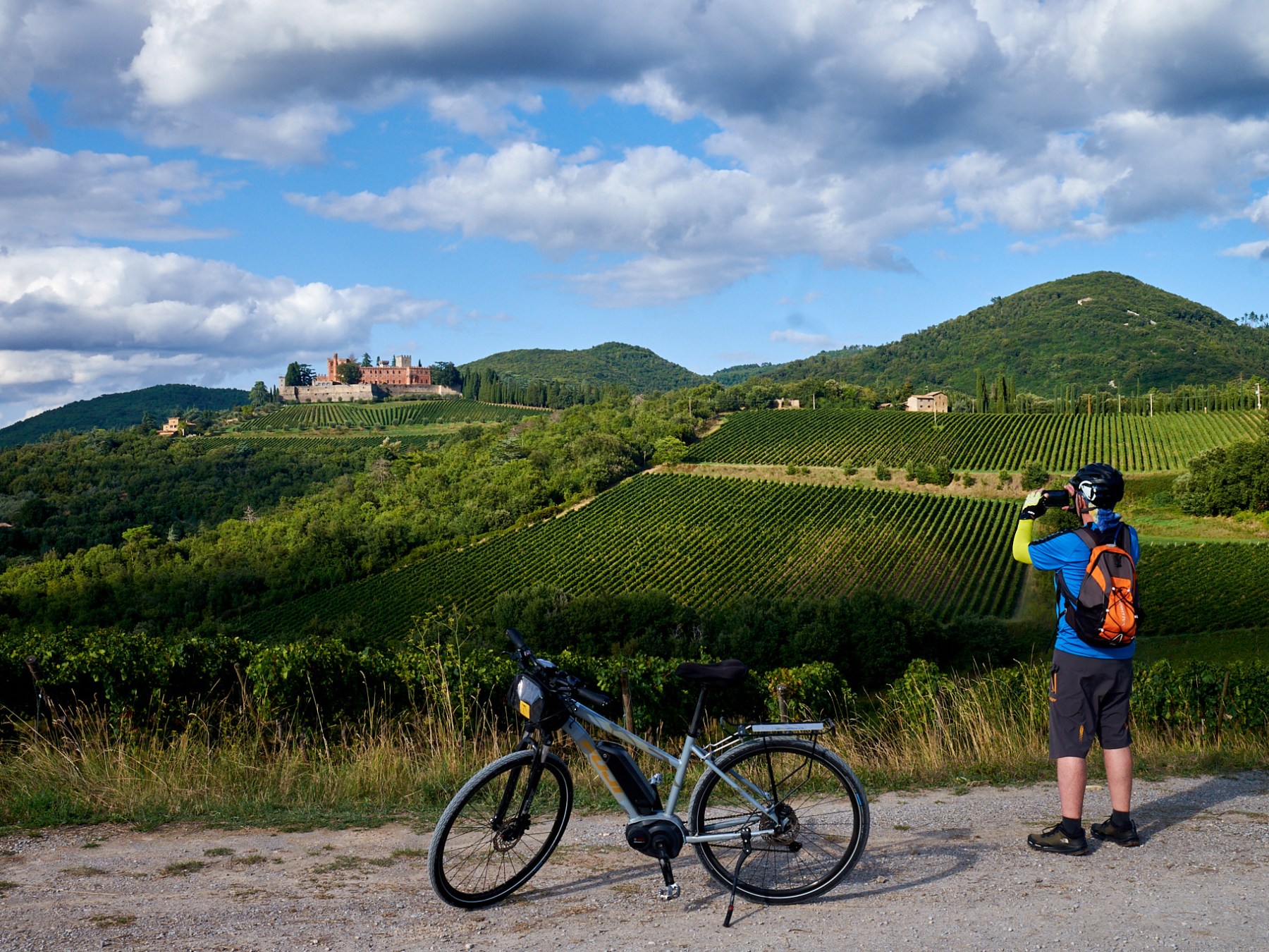 Cyclist photographing scenic vineyard hills and distant castle under cloudy sky.