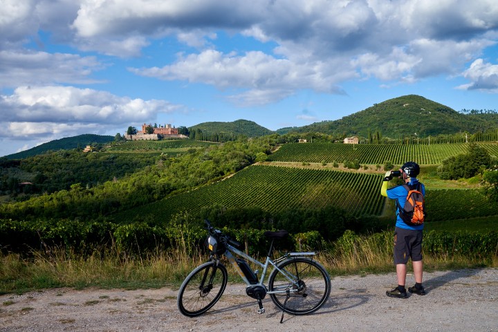 Cyclist photographing scenic vineyard hills and distant castle under cloudy sky.