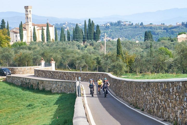 Cyclicsts riding on a scenic path with a stone wall, church, and hills in the background.