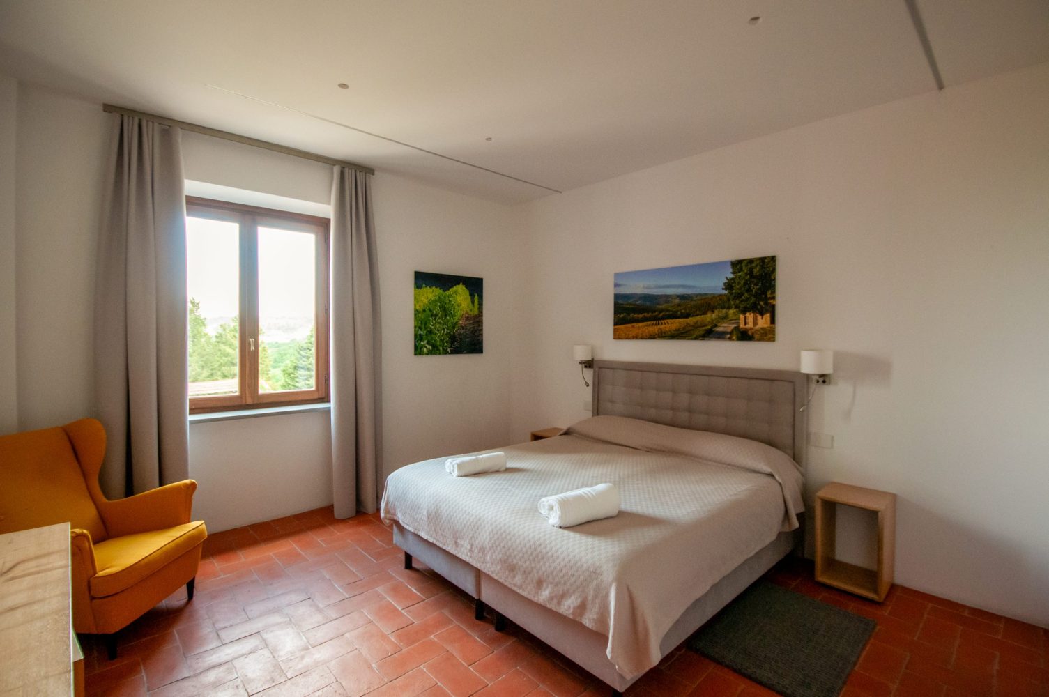 Simple bedroom with double bed, two pictures, window with curtains, and an orange armchair.
