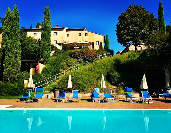 Italian villa on hill with cypress trees, sun loungers, and a pool in foreground under blue sky.