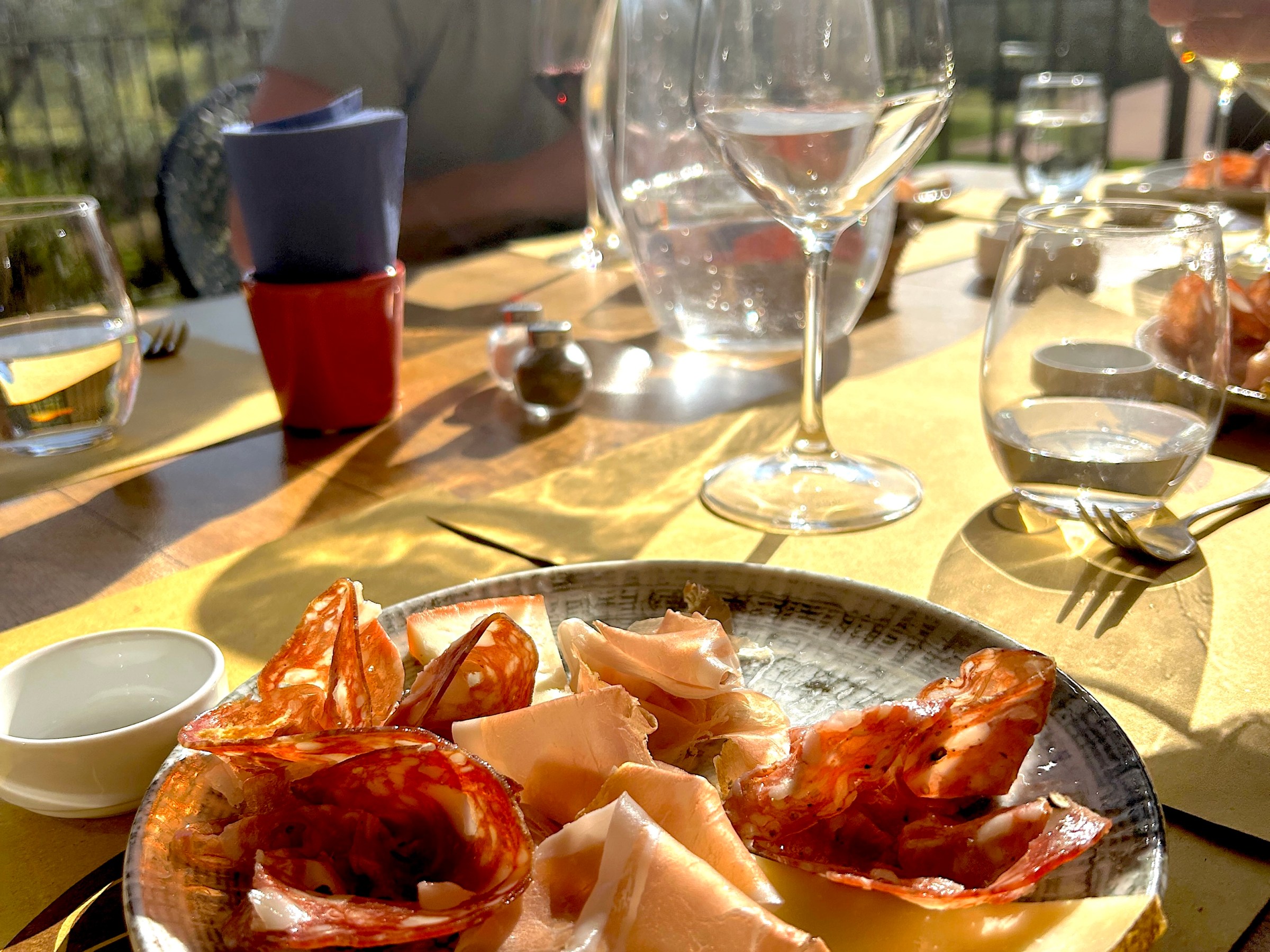 Plate of cured meats and cheese on table with glasses and a blurred person in the background.