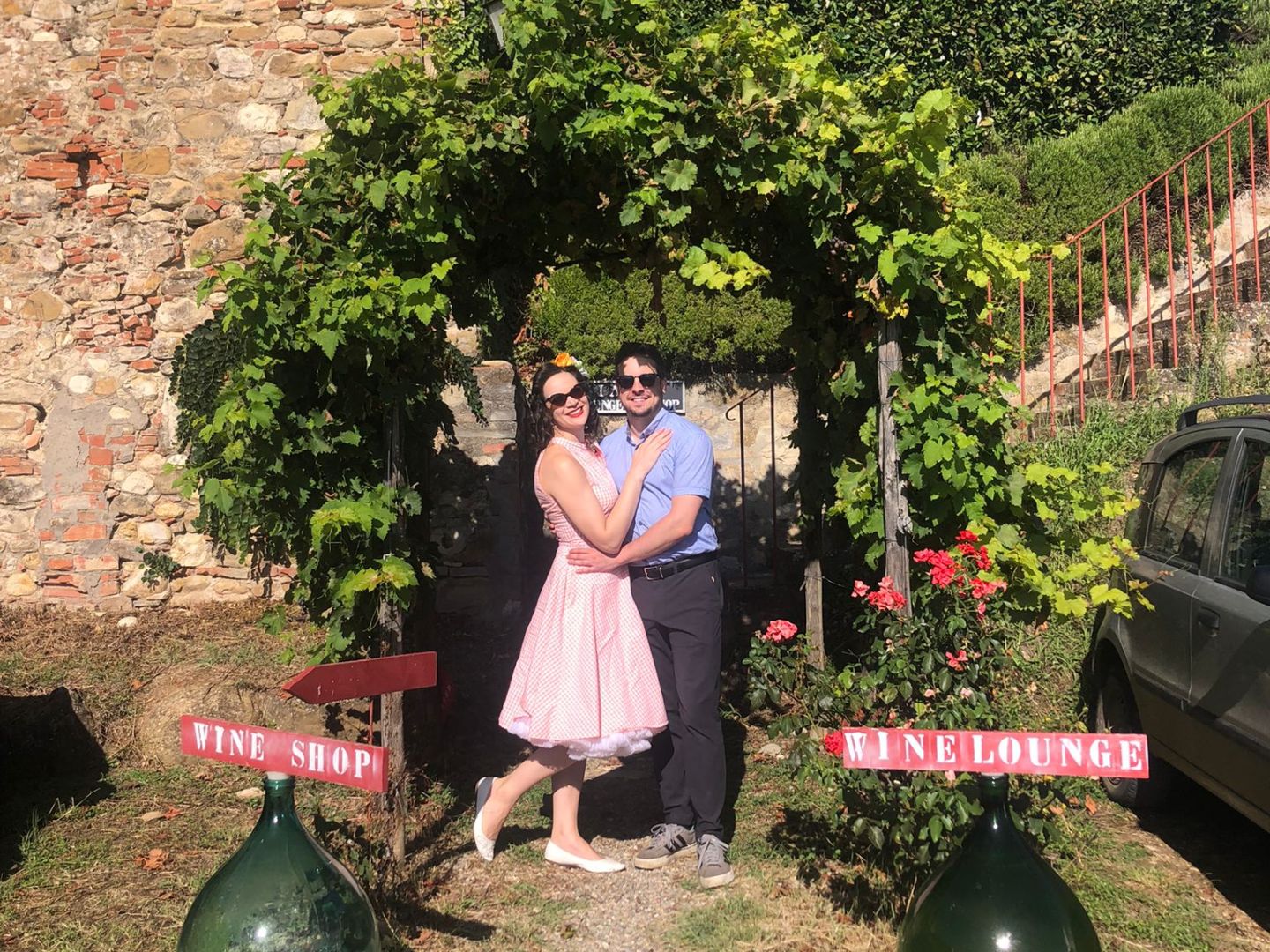 Couple standing under vine archway in front of rustic building with wine signs.