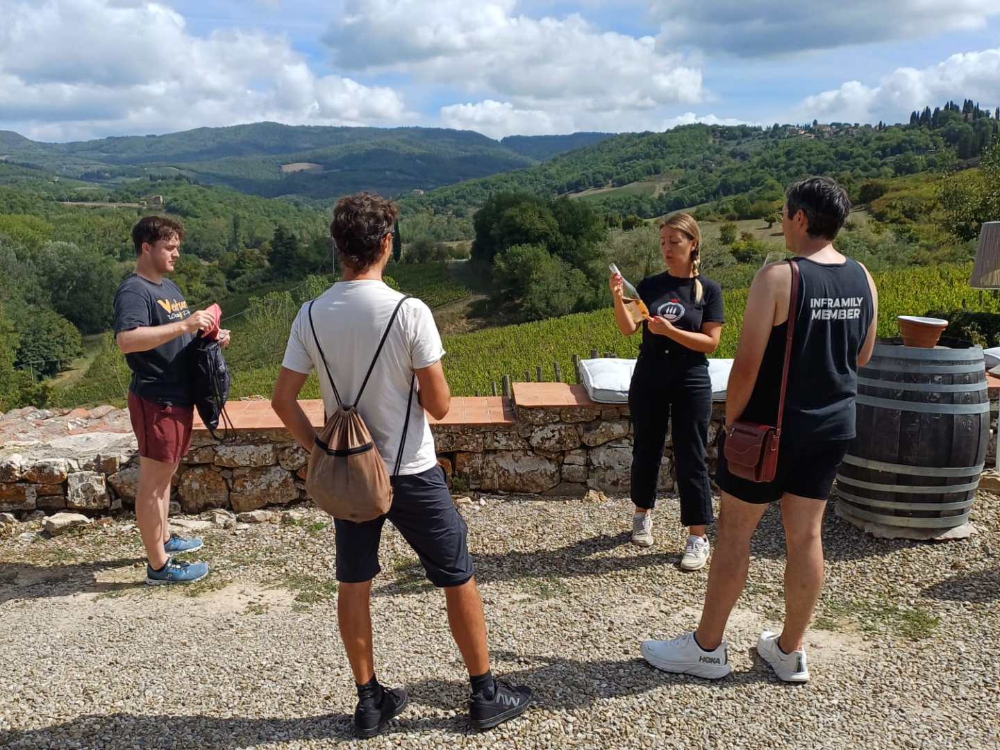 Four people stand on gravel with scenic hills in the background, one holding a wine bottle.