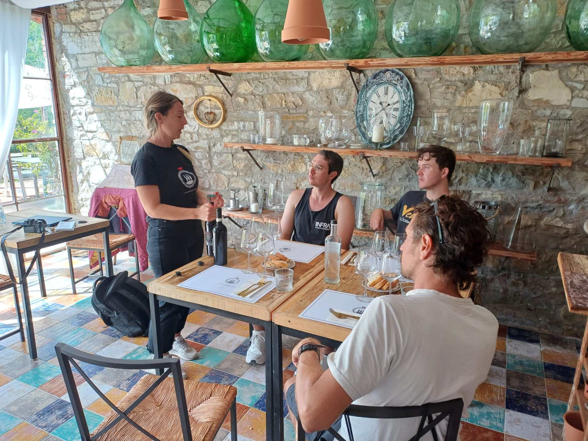 Woman explaining wine to three seated men at a rustic restaurant.