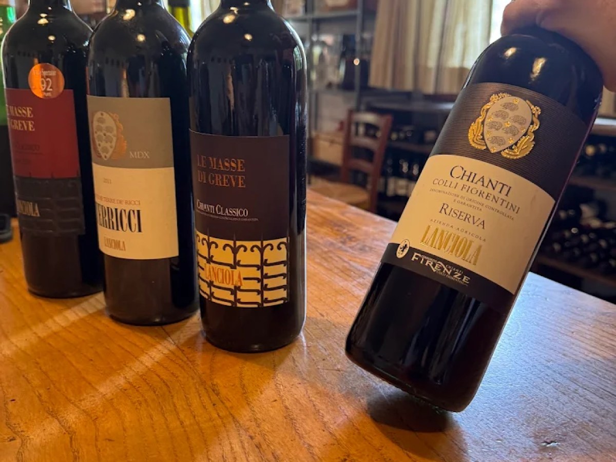 Four wine bottles on a wooden table, with a hand holding one near a window.