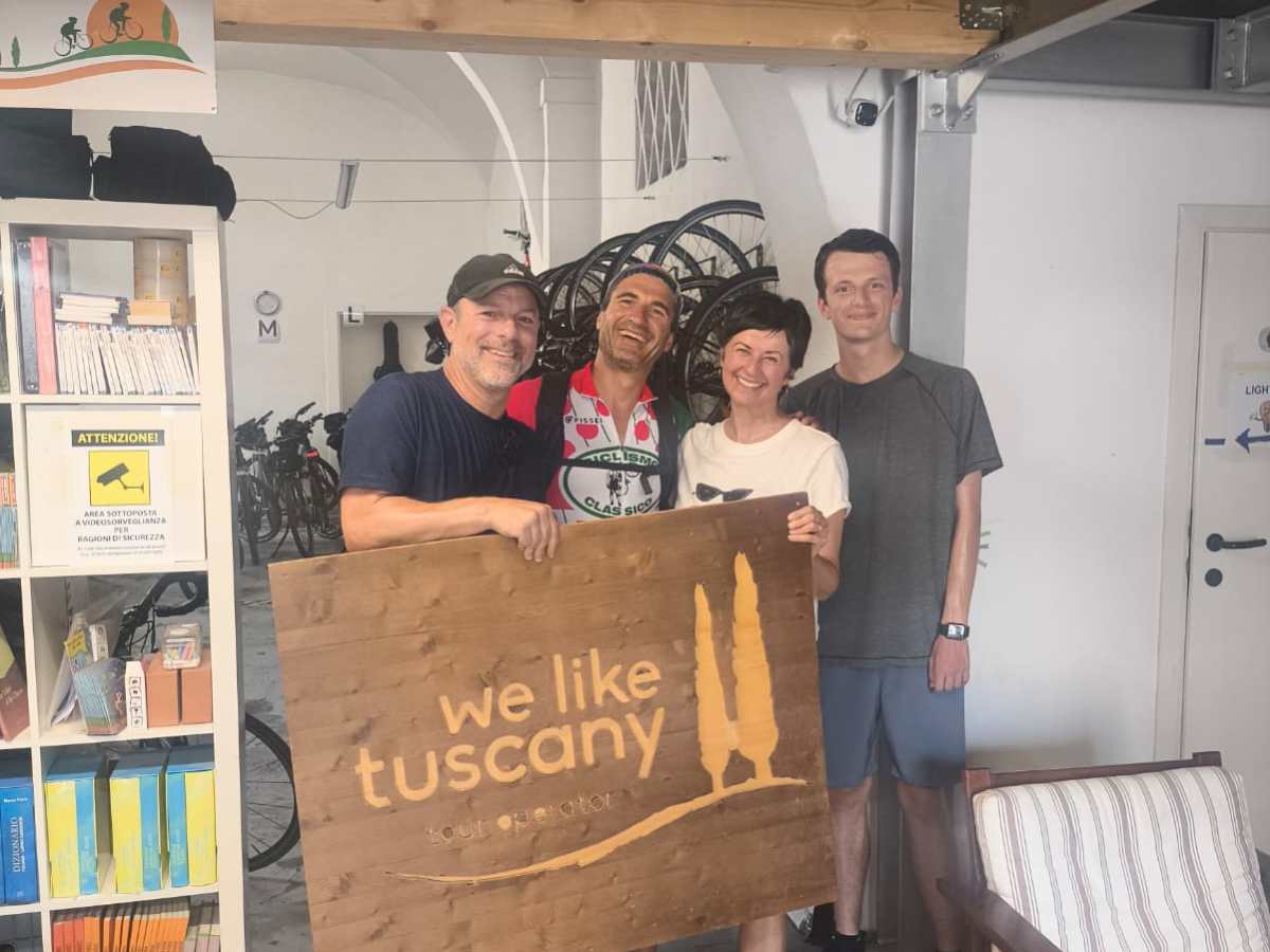 Four people in a room hold a wooden sign saying 'we like tuscany' with books and a couch around them.