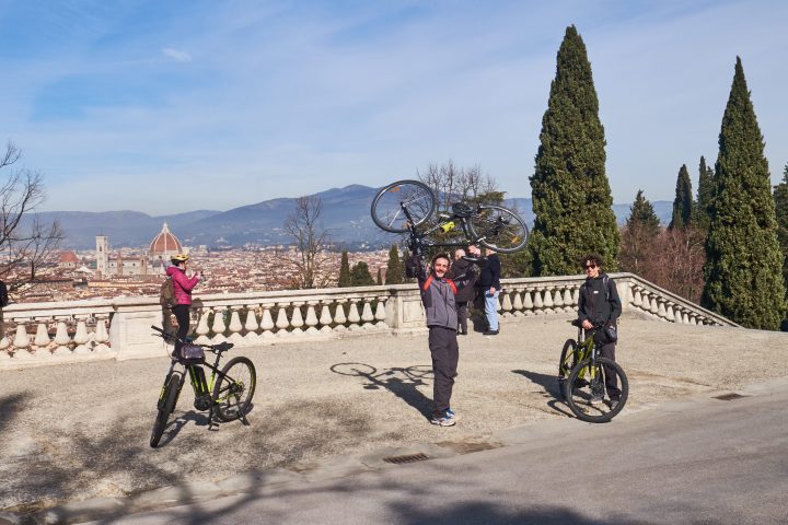 Four people with bicycles enjoying a scenic view from a terrace with trees and a city in the background.