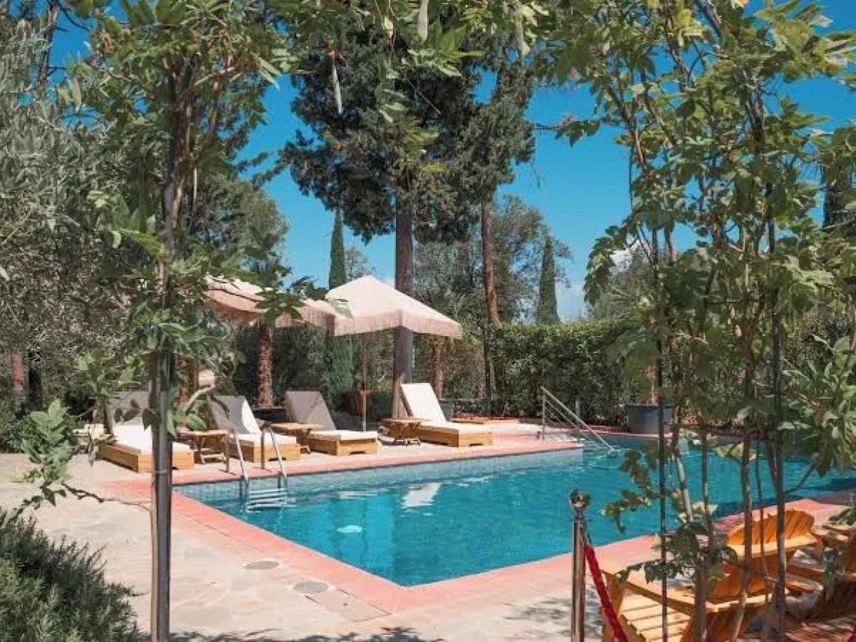 Outdoor pool with sun loungers, umbrellas, and surrounding greenery under a clear blue sky.