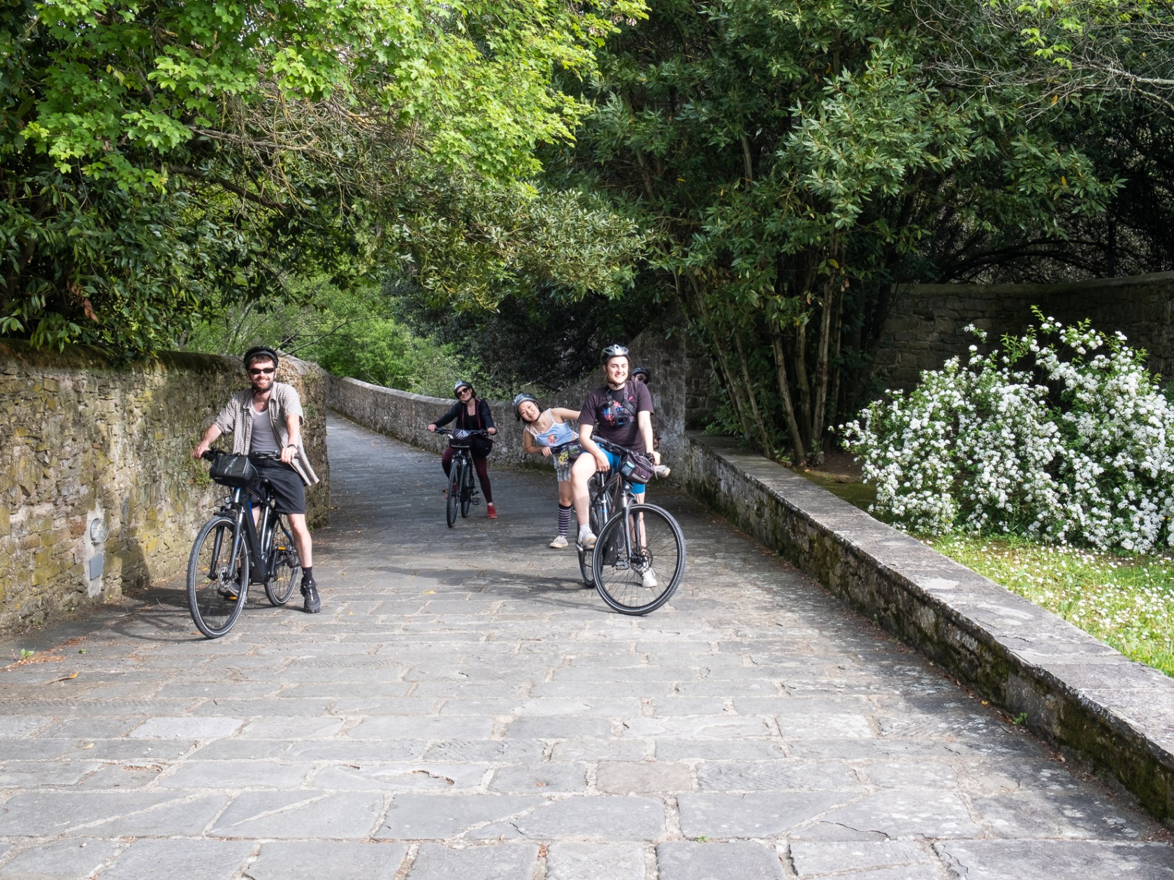 Group of four people cycling on a stone path with trees and a stone wall.