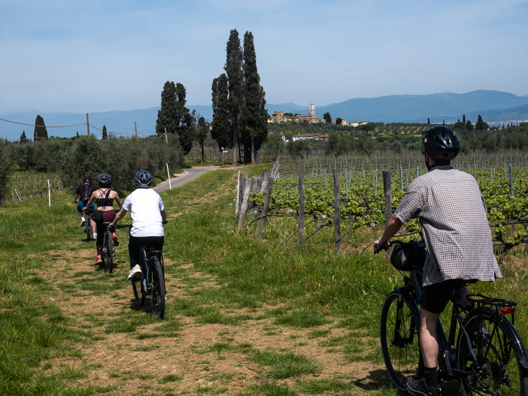 Group cycling along a path through vineyards with a town and mountains in the distance.