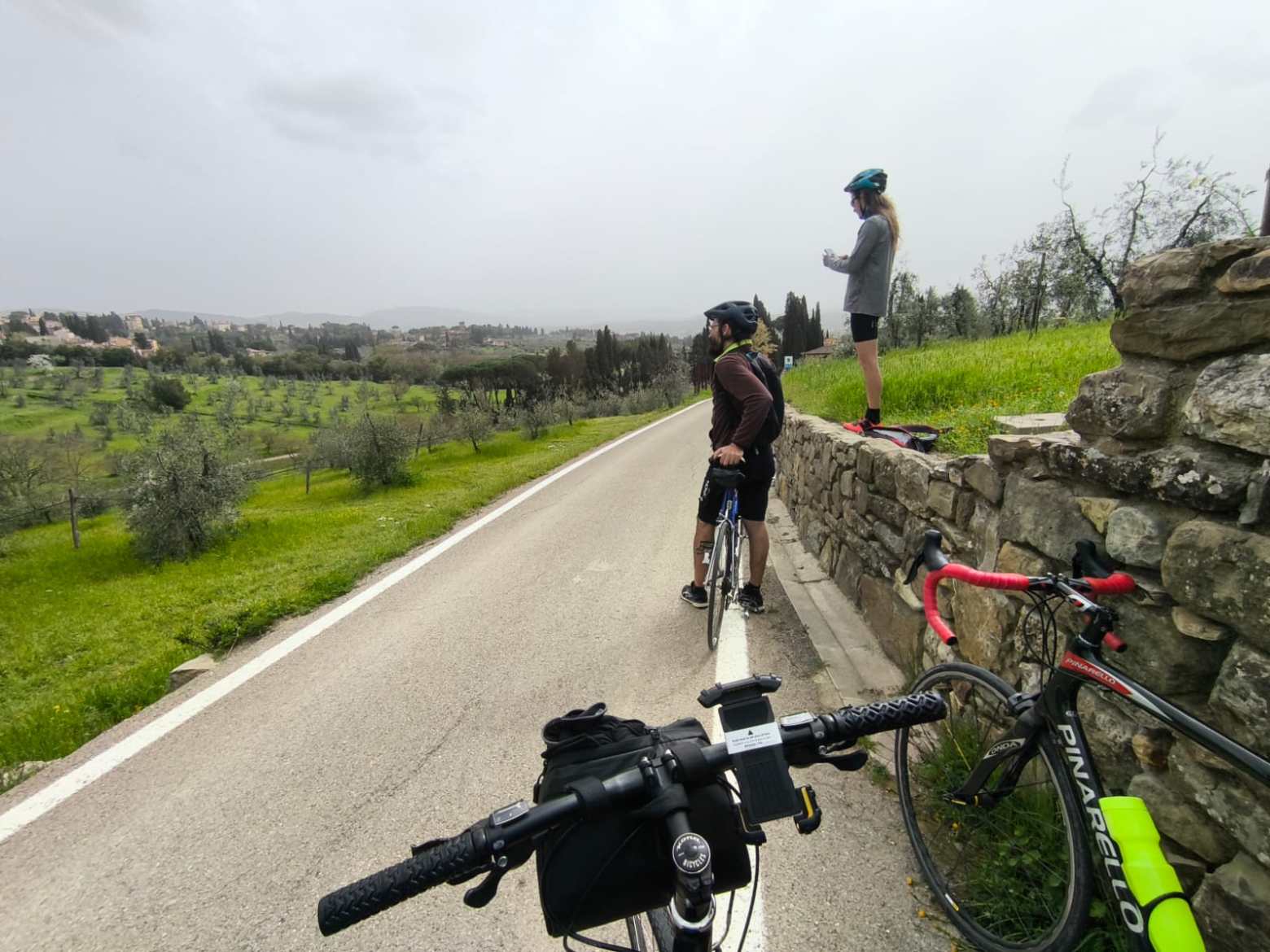 Cyclists paused on a rural road, admiring a scenic landscape with trees and fields.