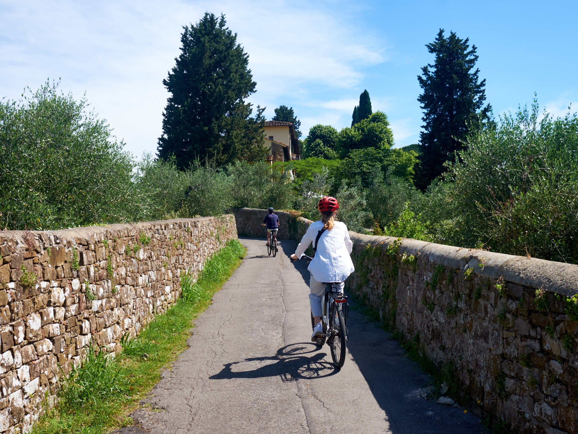 Two people cycling on a narrow path lined with stone walls and trees.