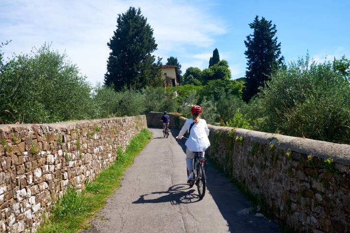 Two people cycling on a narrow path lined with stone walls and trees.
