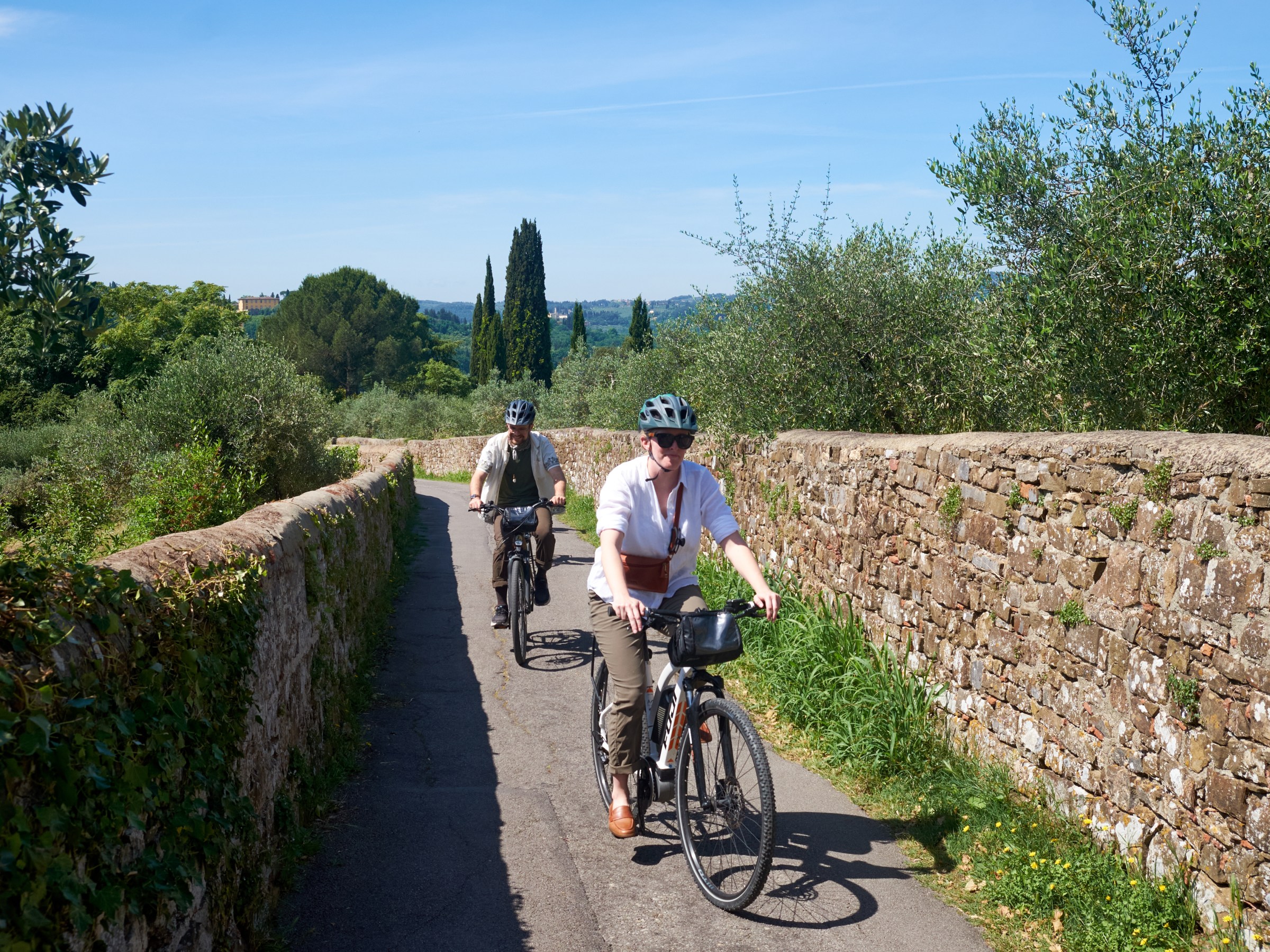 Two people riding bicycles on a narrow country path with stone walls and greenery.