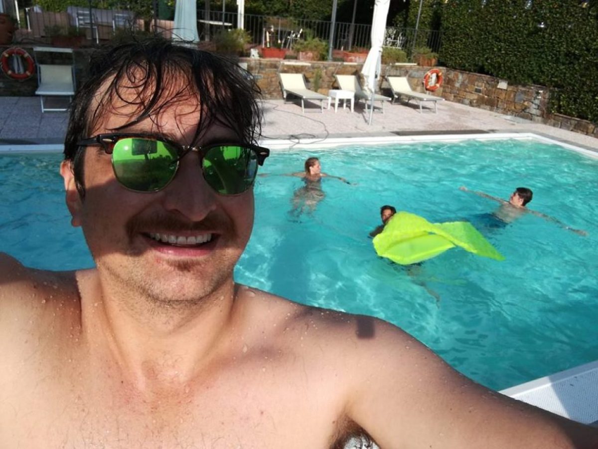 Man in sunglasses taking selfie at a pool with three people swimming in background.