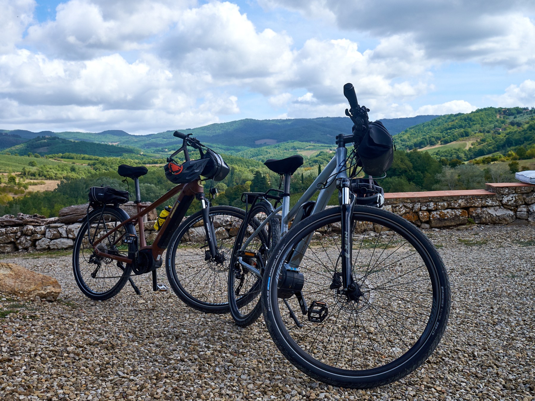 Two bicycles parked on gravel with a scenic hilly landscape in the background under cloudy skies.