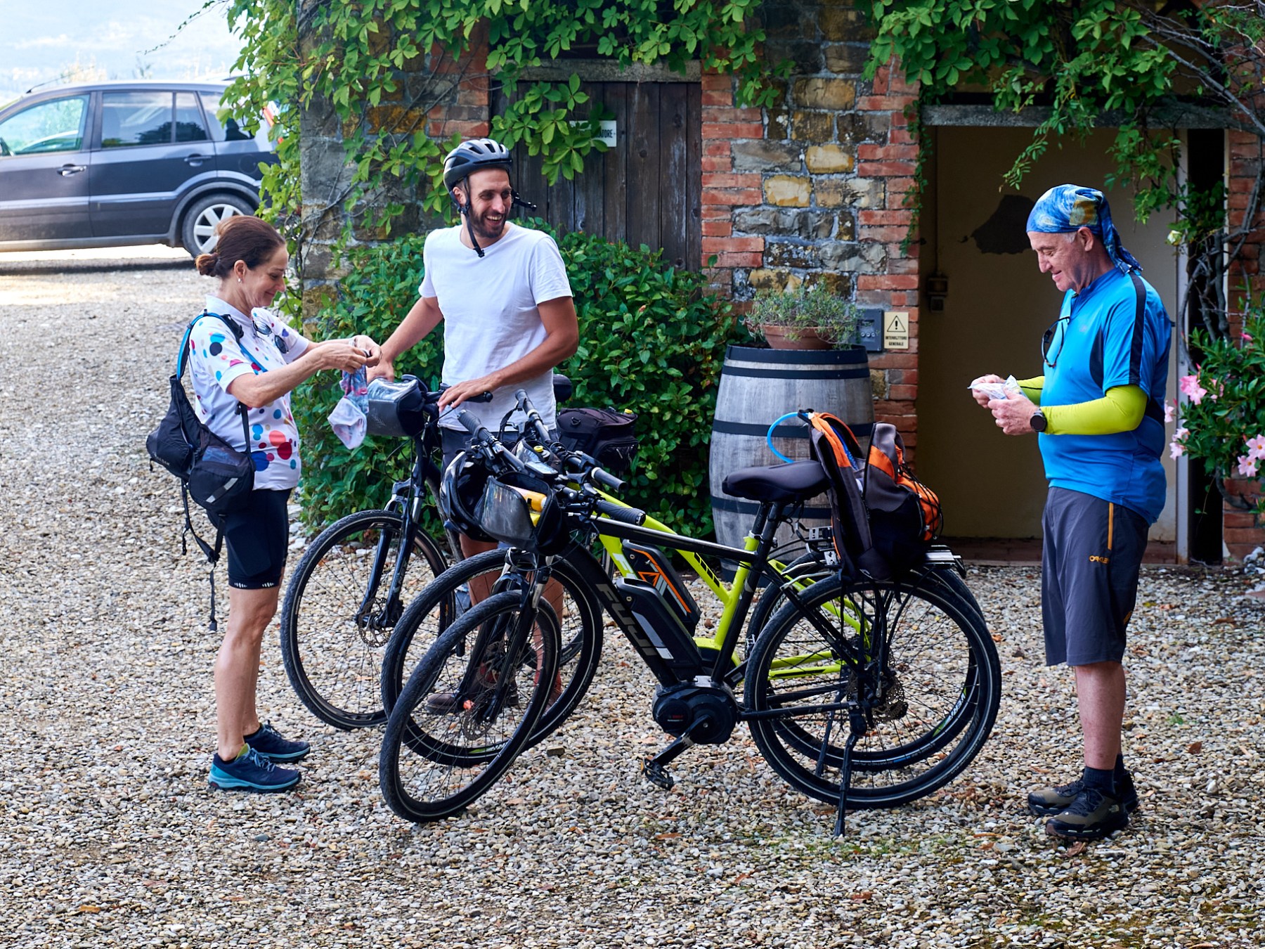 Three bicyclists prepare their bikes near a stone building with plants around.