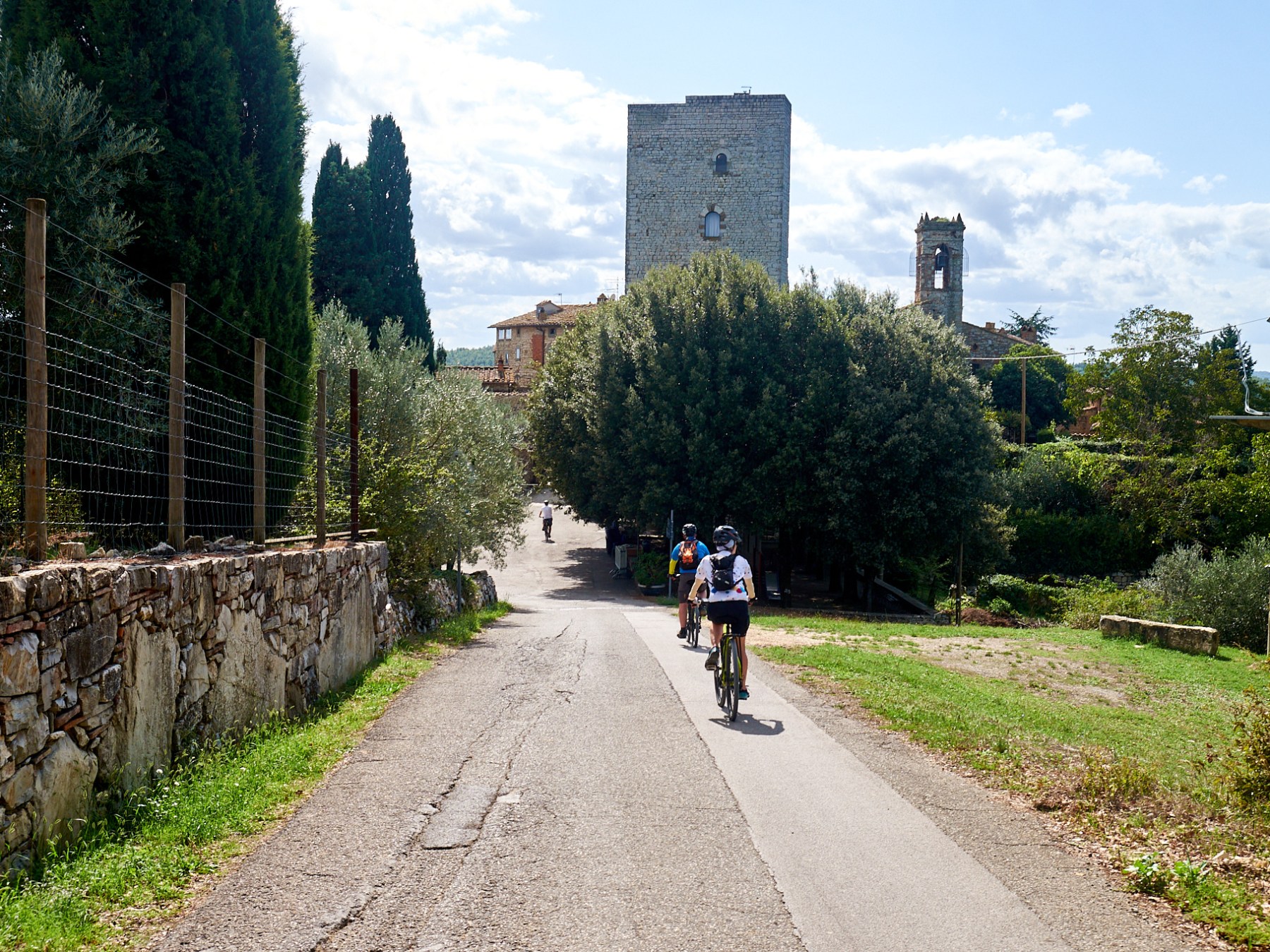 Cyclists on a rural path with stone wall, trees, and a tower in the background.