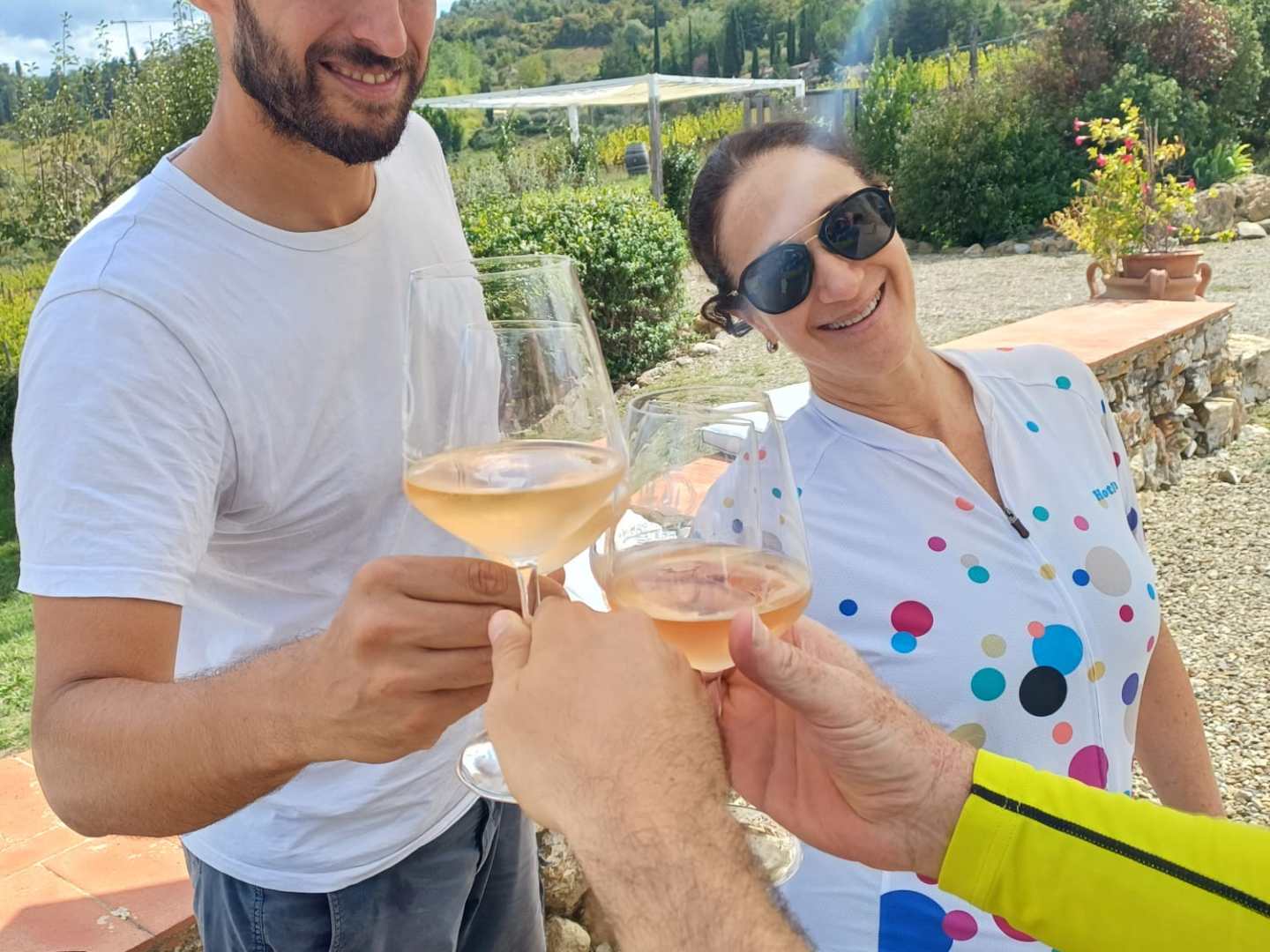 Three people toast with wine glasses under a sunny patio with umbrellas and a scenic background.
