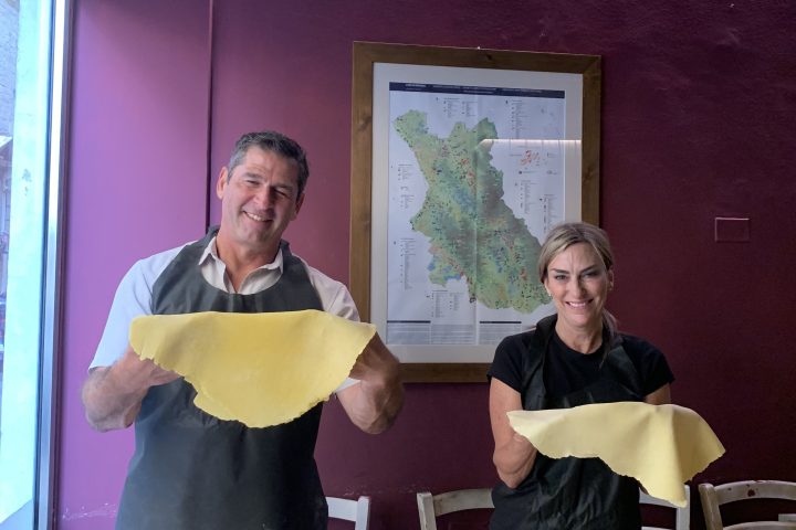 Two people holding pasta sheets in a kitchen with dark red walls.
