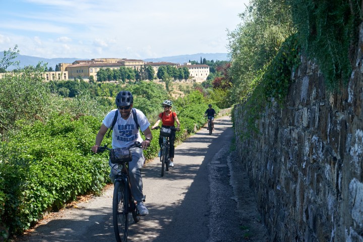 Three cyclists riding along a path with greenery and hillside buildings in the background.