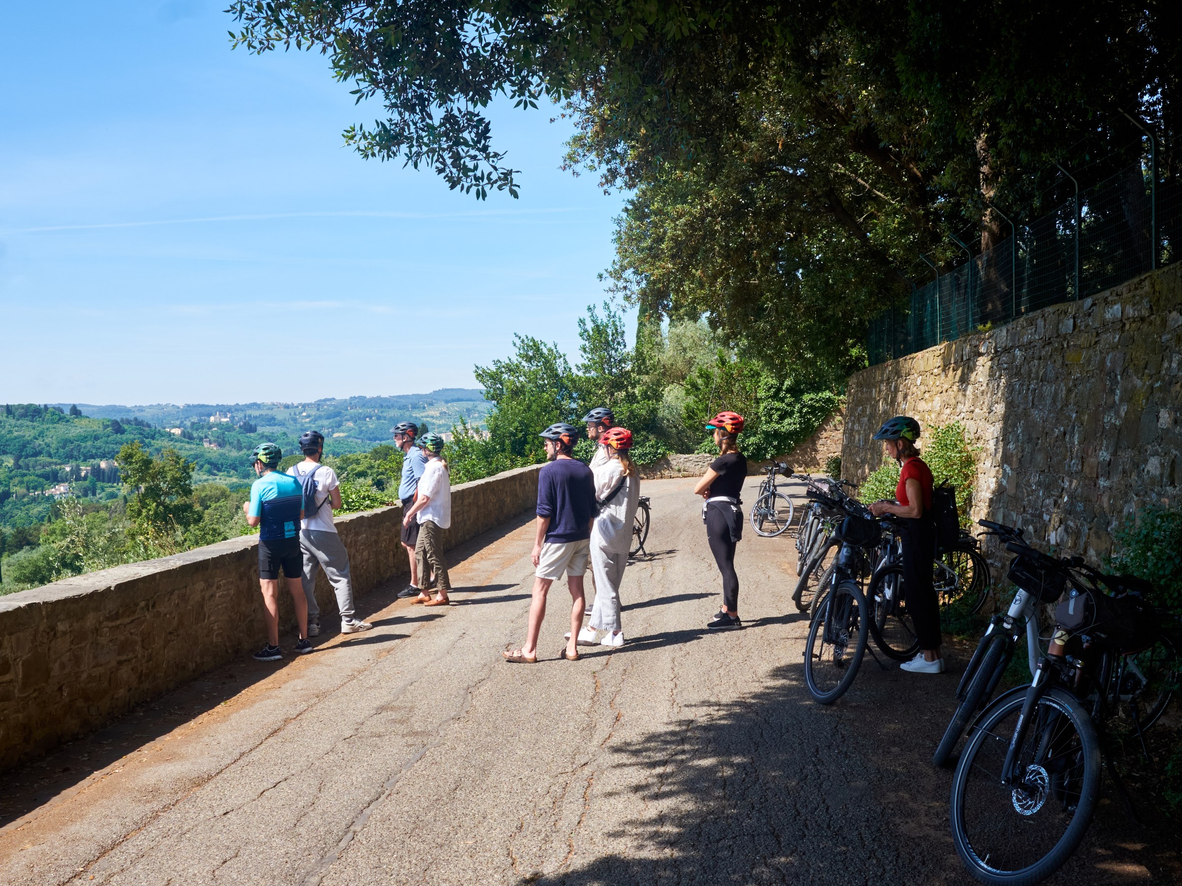 Cyclists resting on a paved road overlooking a scenic valley, with bikes parked beside a stone wall.