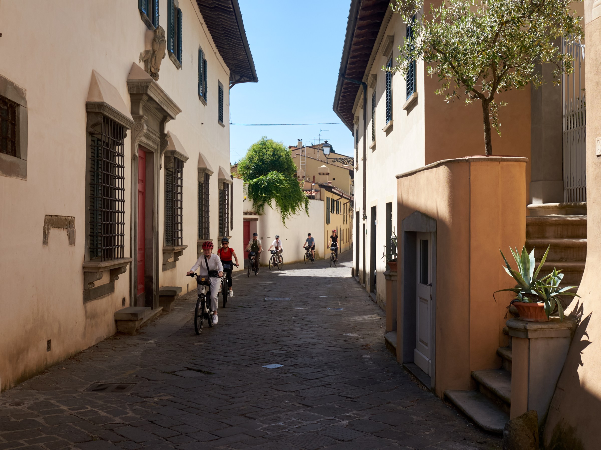 Cyclists ride down a narrow, sunny alley flanked by old buildings and potted plants.