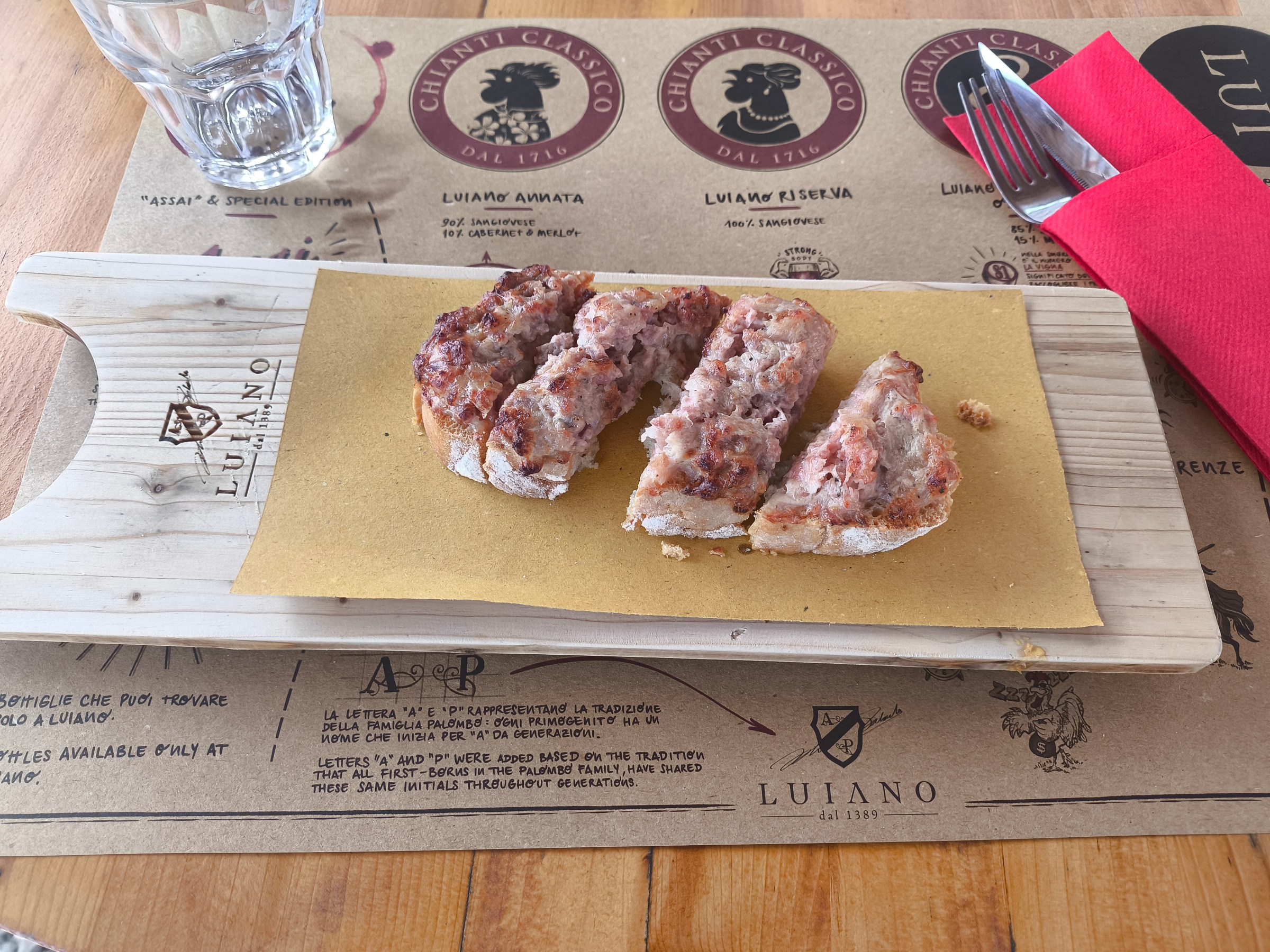 Sliced sausage on bread served on a wooden board with a glass.