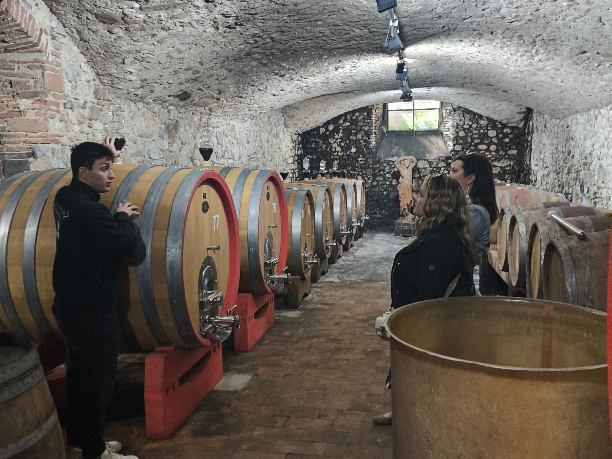 People in a stone cellar with large wine barrels lining the walls.