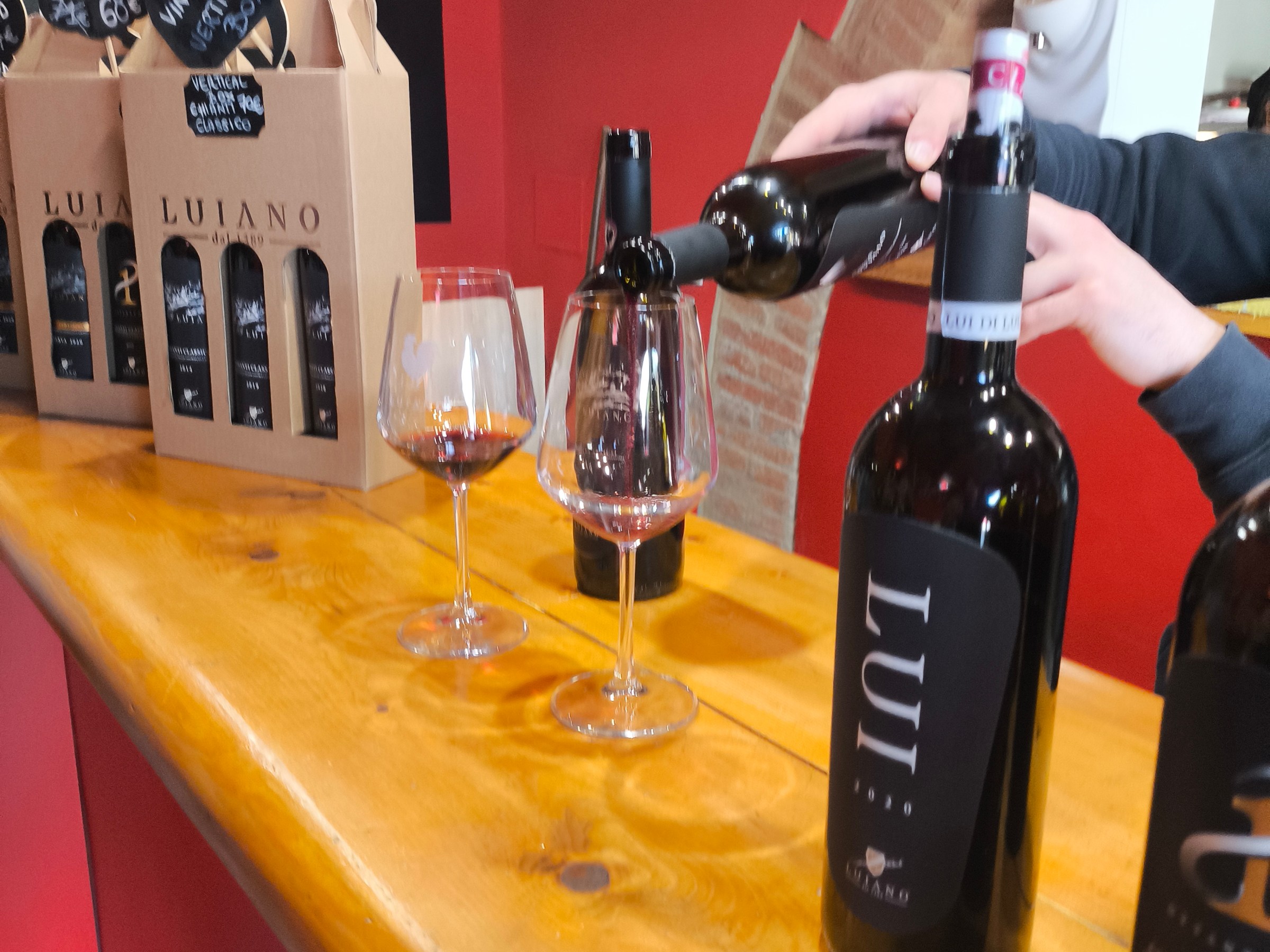 Person pouring red wine into a glass at a wooden counter with wine bottles.
