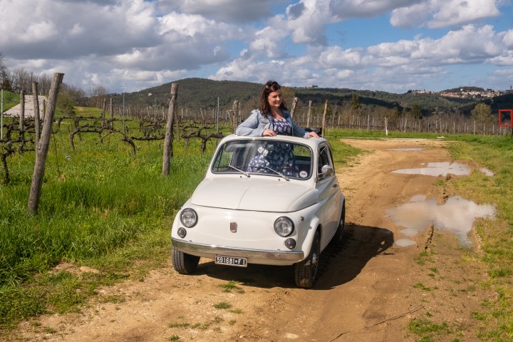 Person standing in a small white car on a dirt road in a vineyard.
